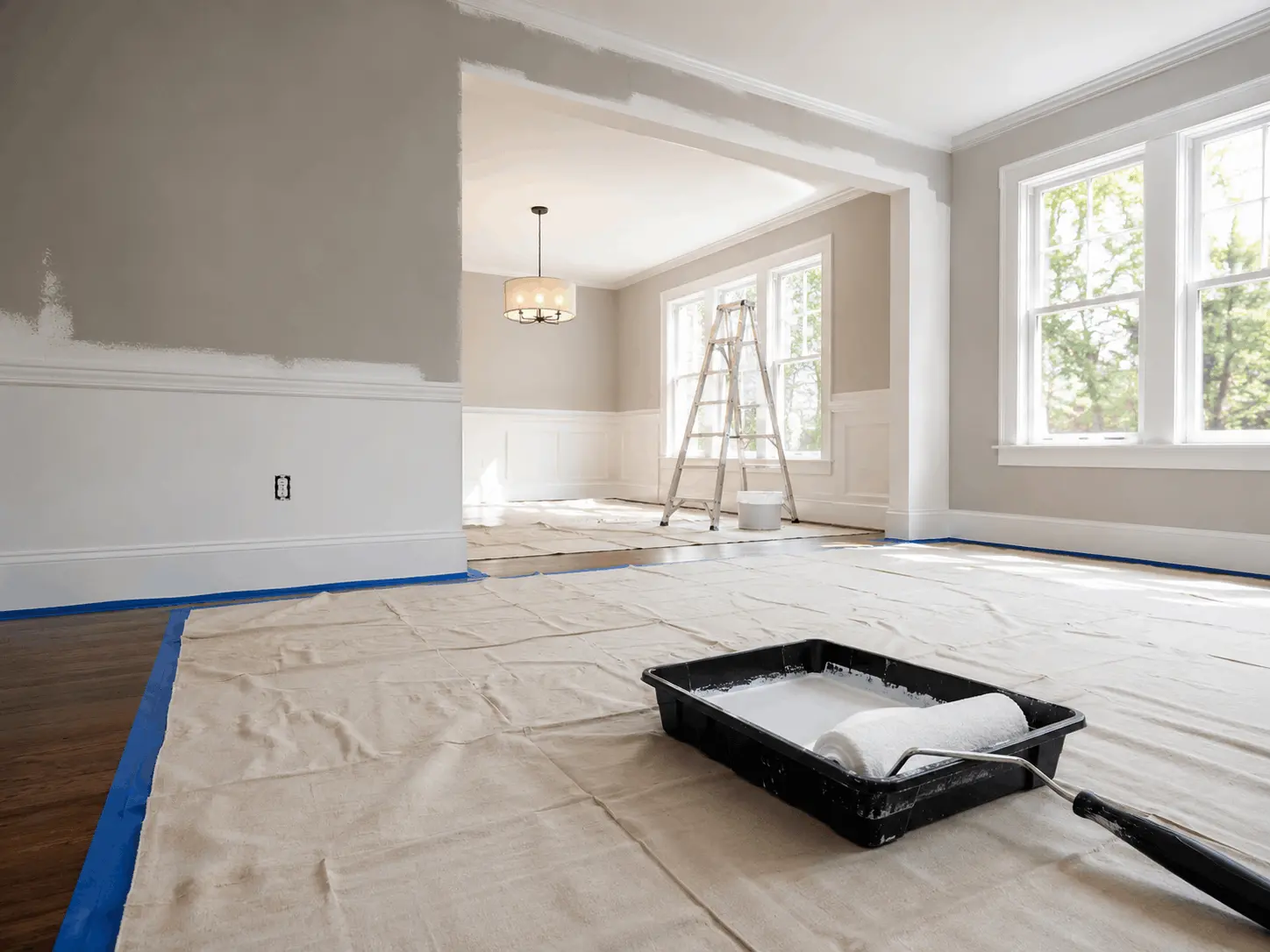 A wide shot of a bright, sunlit room mid-painting, with a canvas drop cloth neatly taped down over hardwood floors, a paint tray, and a roller in the foreground